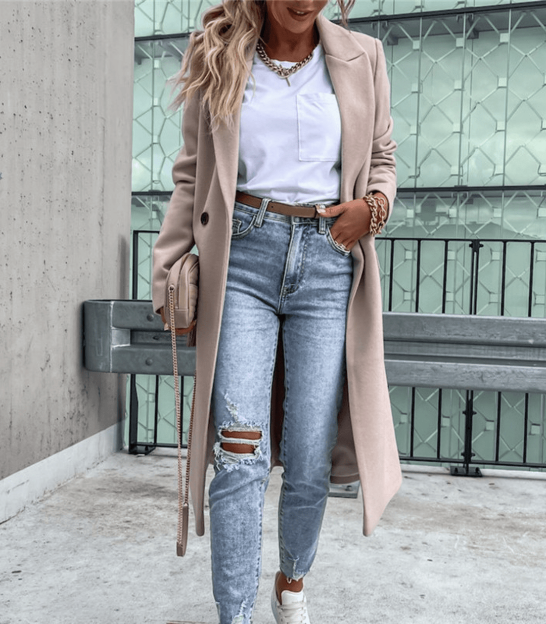 Femme en manteau beige, jean déchiré et baskets blanches, posant devant une structure moderne. Mode urbaine chic, style décontracté élégant.