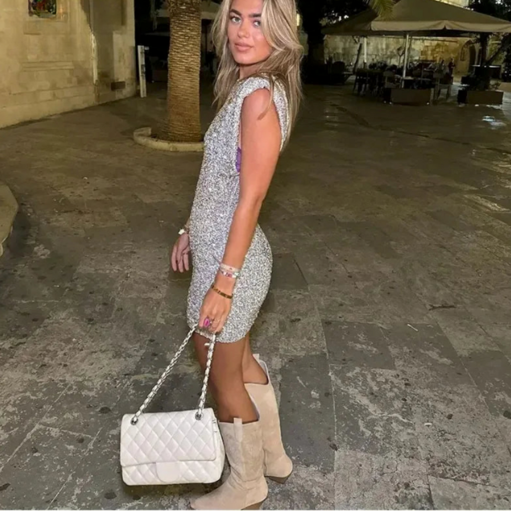 Jeune femme en robe pailletée argentée, portant un sac à main blanc et des bottes beiges, posant sur une place pavée en soirée.
