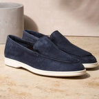 Mocassins en daim bleu marine pour homme, semelle blanche, posés sur un sol beige. Chaussures élégantes et confortables, style décontracté.