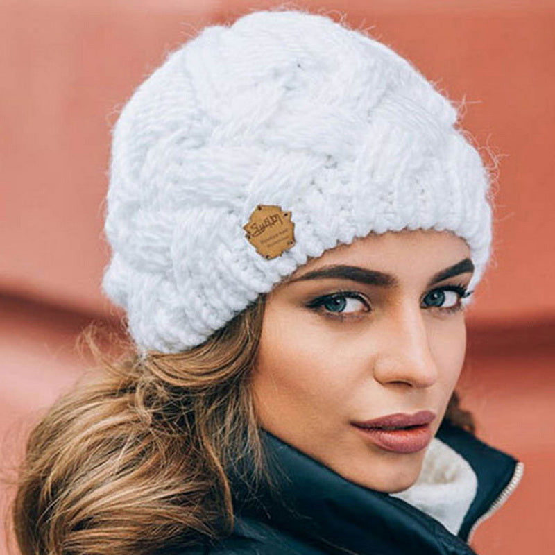 Femme portant un bonnet en laine blanc tricoté, étiquette en cuir, fond flou orange. Mode hiver, accessoires chauds, style élégant.