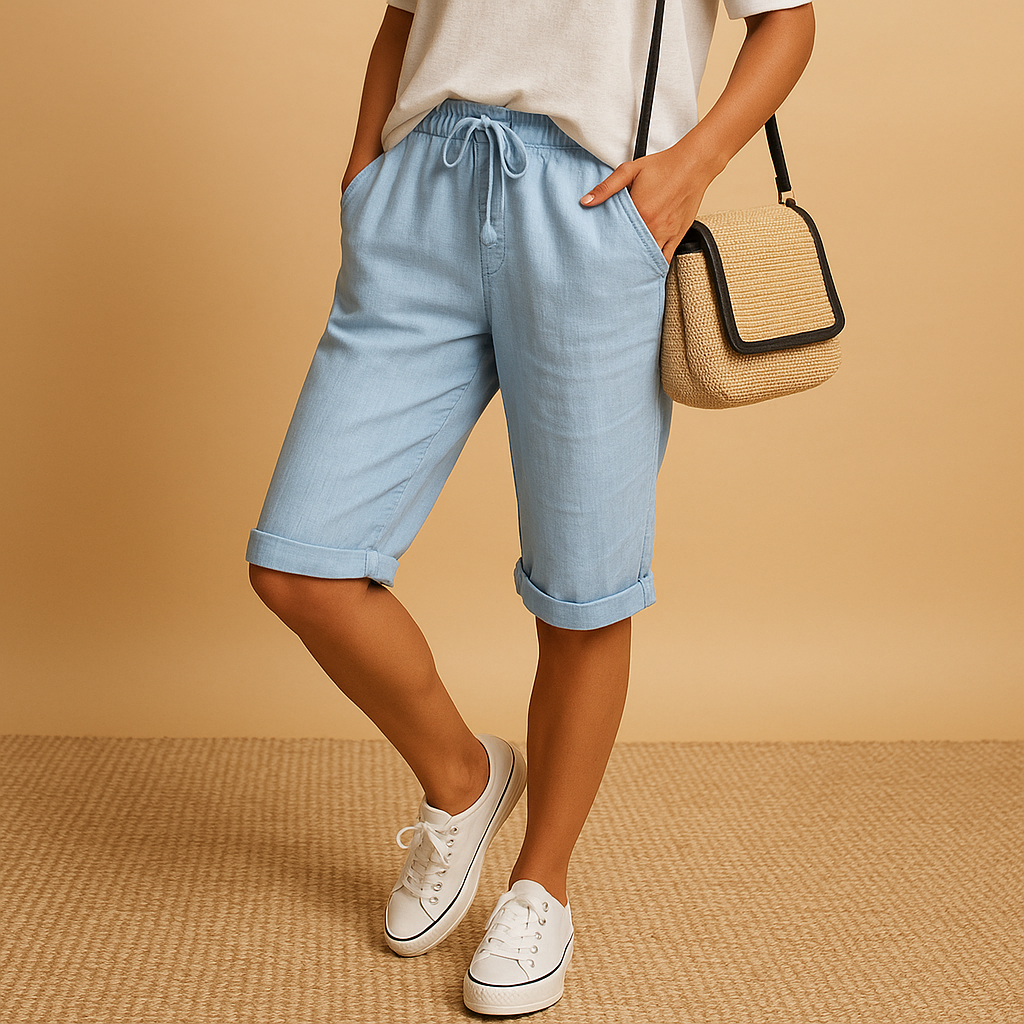 Femme portant un short bleu clair en lin, t-shirt blanc, baskets blanches et sac à main en paille, posant sur fond beige. Mode estivale décontractée.
