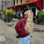 Femme blonde en veste rouge devant un café avec terrasse, cheveux longs ondulés, ambiance urbaine, journée d'automne, Amsterdam.