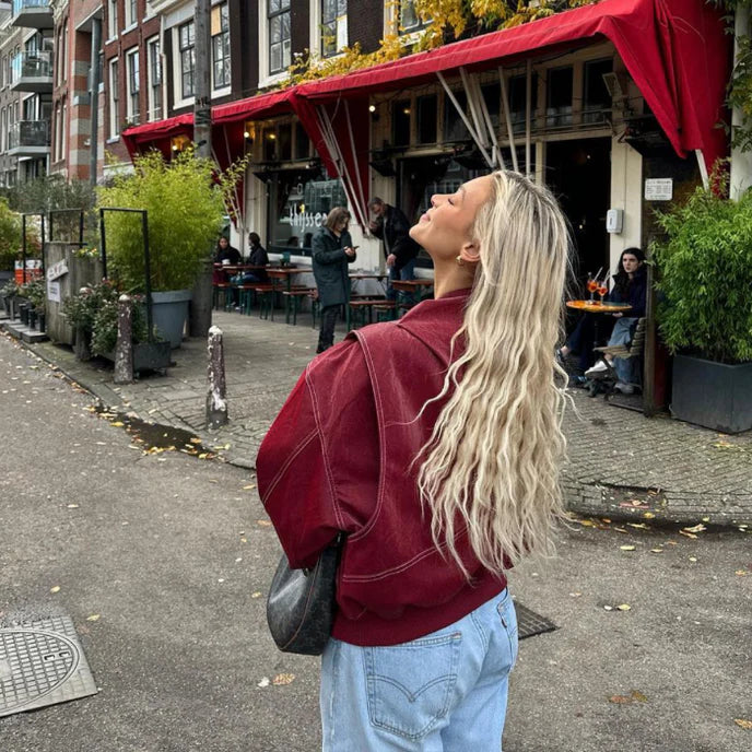 Femme blonde en veste rouge devant un café avec terrasse, cheveux longs ondulés, ambiance urbaine, journée d'automne, Amsterdam.