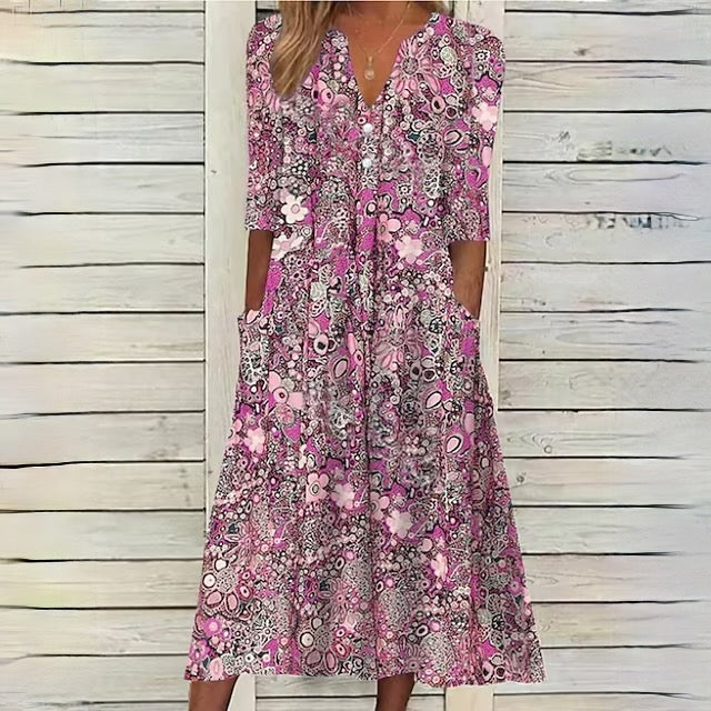 Robe longue bohème rose à motifs floraux, manches trois-quarts, col en V, poches latérales, fond en bois blanc. Mode féminine estivale.