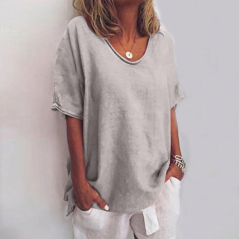 Femme portant un t-shirt ample gris, col rond, manches courtes, avec un collier et un bracelet, posant contre un fond neutre. Mode décontractée élégante.