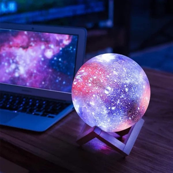 Lampe galaxie LED colorée sur support en bois, posée à côté d'un ordinateur portable, créant une ambiance cosmique et lumineuse dans une pièce sombre.