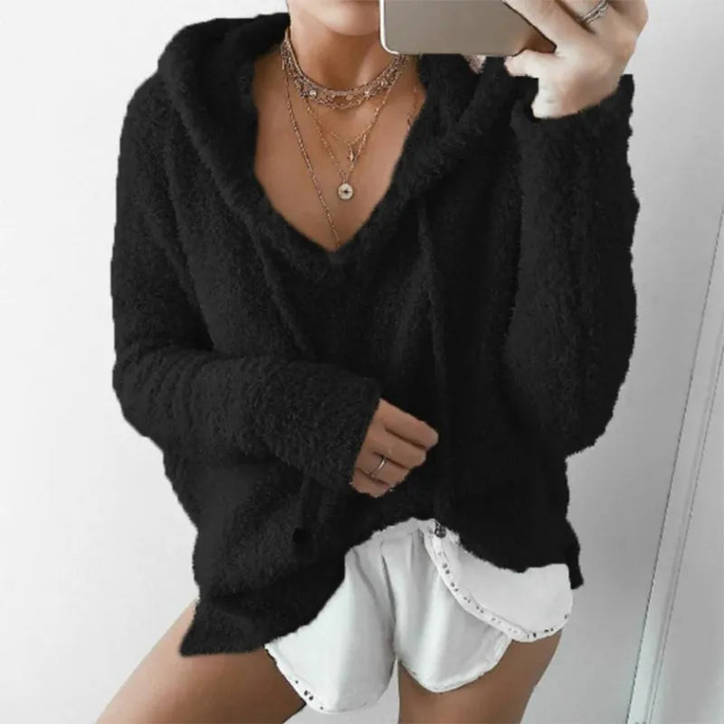 Femme portant un pull noir à capuche, col en V, avec des shorts blancs. Style décontracté, mode automne-hiver, tendance vêtements confortables.