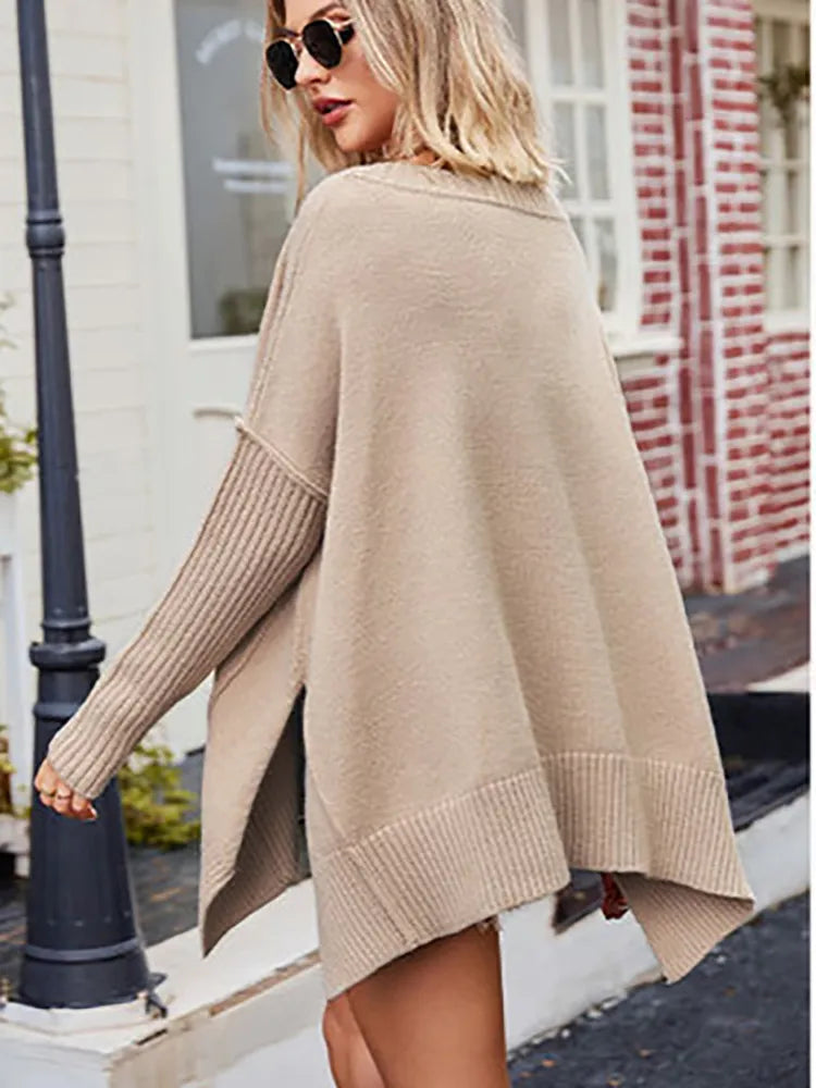 Femme portant un pull beige oversize, manches longues, devant une maison en briques rouges. Mode automne, style décontracté, lunettes de soleil.