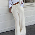 Femme en pantalon large blanc, haut court blanc, tenant un gobelet à café, posant contre un mur extérieur. Mode décontractée, style urbain.
