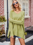 Femme portant un pull vert ample, manches longues, col en V, avec des lunettes de soleil, devant une maison en briques rouges. Mode automne décontractée.