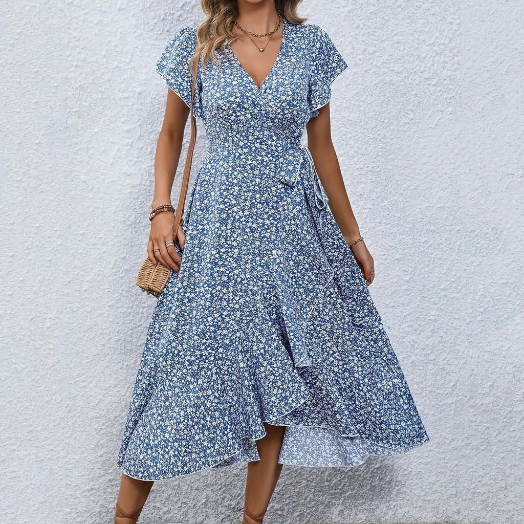 Robe d'été bleue à motifs floraux, manches courtes, ceinture nouée, portée par une femme devant un mur blanc. Mode féminine élégante et décontractée.