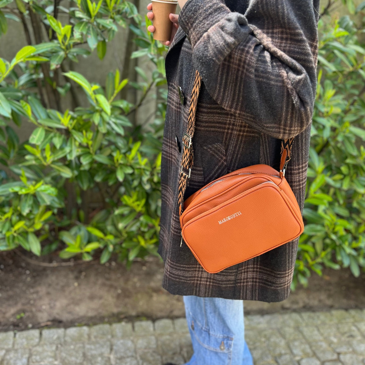 Personne portant un manteau à carreaux, tenant un café, avec un sac à bandoulière orange Balibeyti. Fond de verdure. Mode automne décontractée.