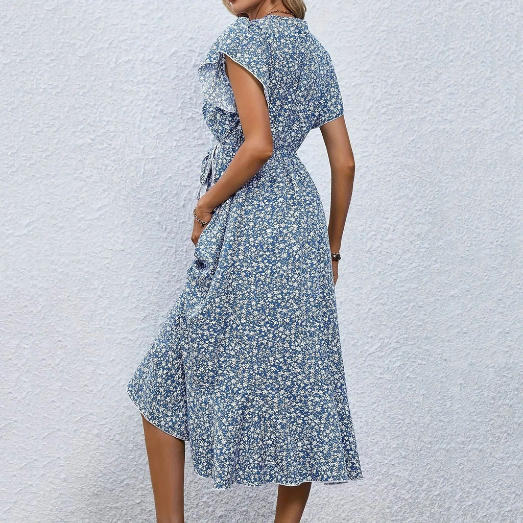 Robe d'été bleue à motifs floraux, manches courtes, ceinture à la taille, portée par une femme sur fond de mur blanc. Mode féminine élégante.