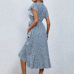 Robe d'été bleue à motifs floraux, manches courtes, ceinture à la taille, portée par une femme sur fond de mur blanc. Mode féminine élégante.