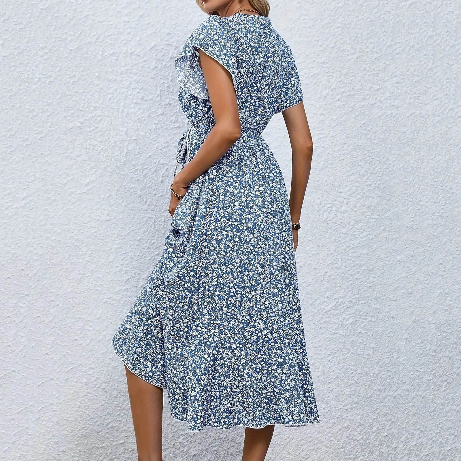 Robe d'été bleue à motifs floraux, manches courtes, ceinture à la taille, portée par une femme sur fond de mur blanc. Mode féminine élégante.