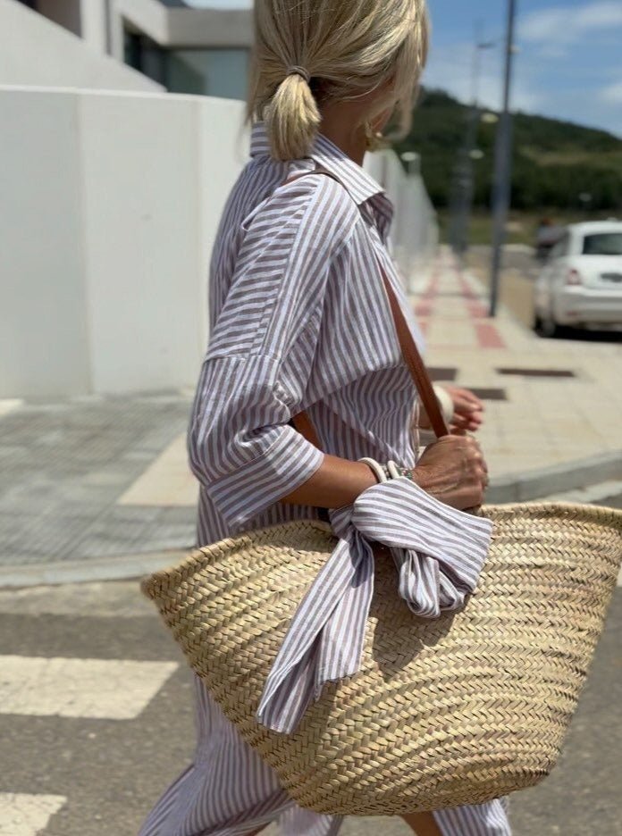 Femme en chemise rayée, portant un grand sac en osier, marchant en extérieur. Mode estivale, style décontracté, accessoire tendance.