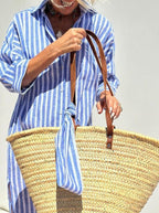 Femme en robe à rayures bleues portant un grand sac en osier avec anses en cuir, accessoire estival tendance, mode plage chic.