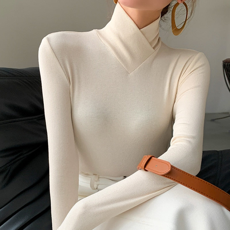 Femme portant un pull col roulé beige, manches longues, avec des boucles d'oreilles dorées et un sac en cuir marron. Mode élégante et minimaliste.