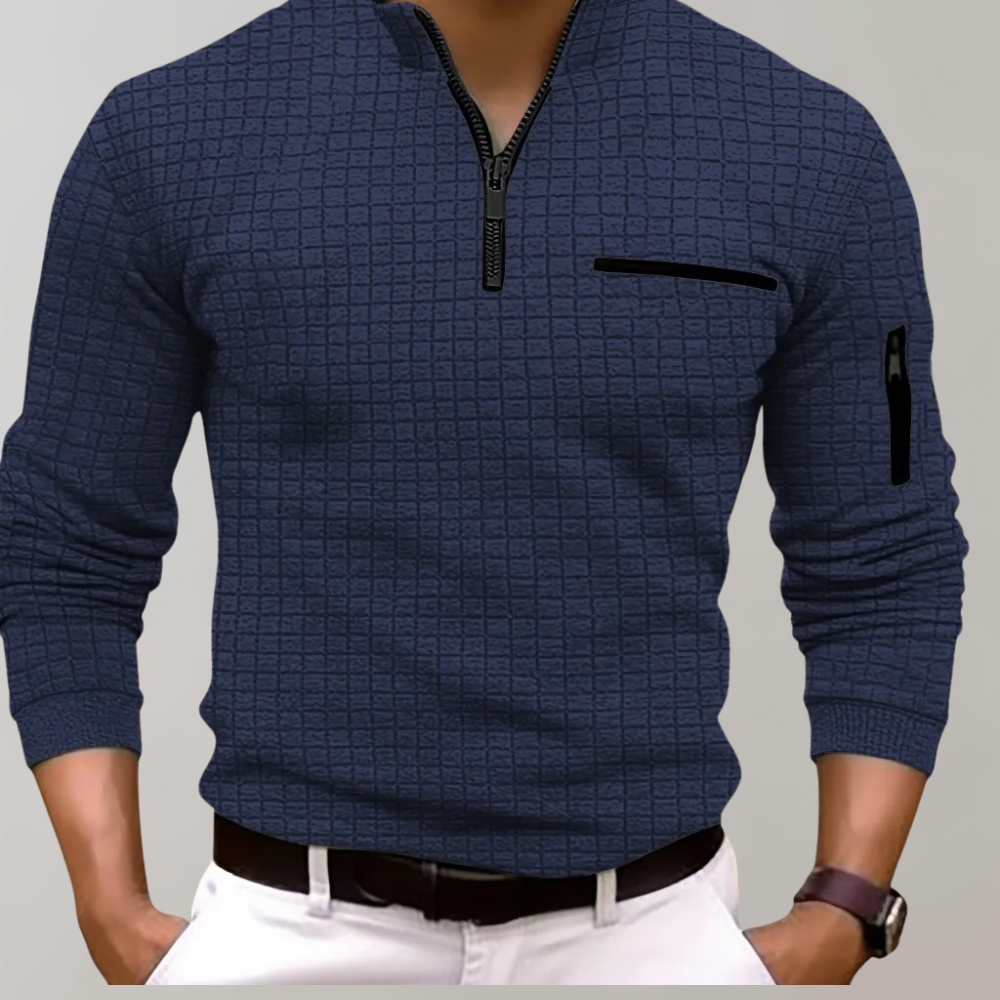 Homme portant un pull bleu à col zippé, motif quadrillé, avec poches zippées, manches longues, ceinture marron, pantalon blanc. Mode masculine élégante.