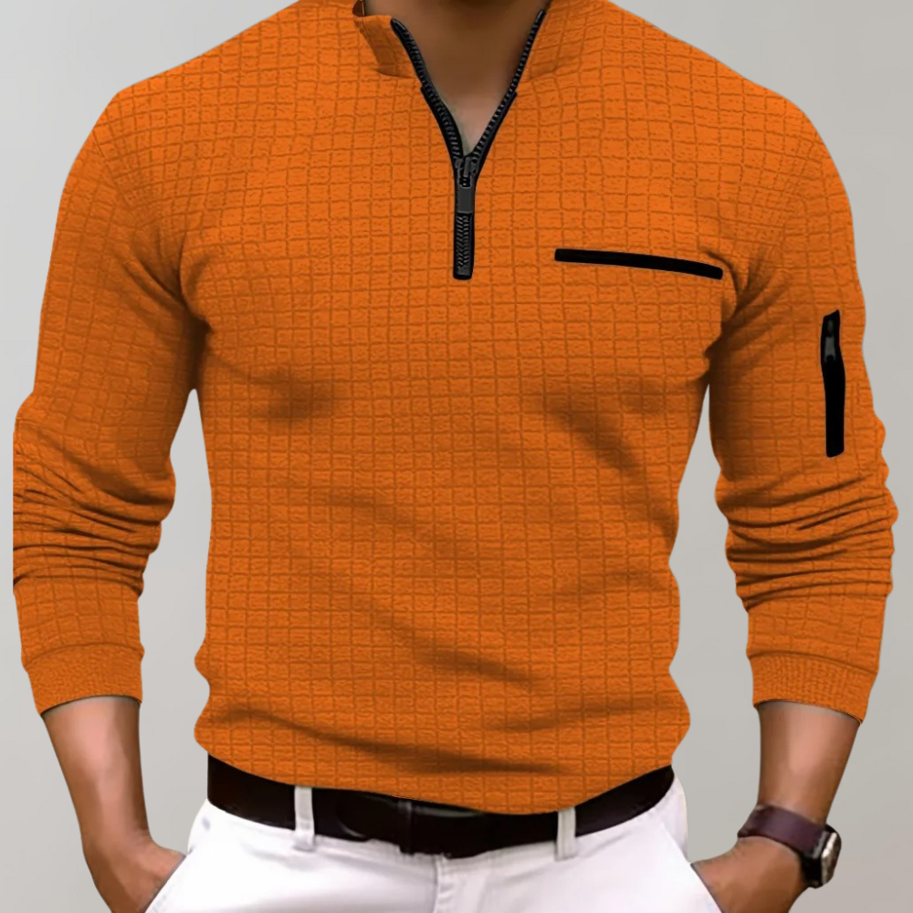 Homme portant un pull orange à col zippé, motif quadrillé, avec poche poitrine et manche, sur fond gris. Mode masculine élégante et moderne.