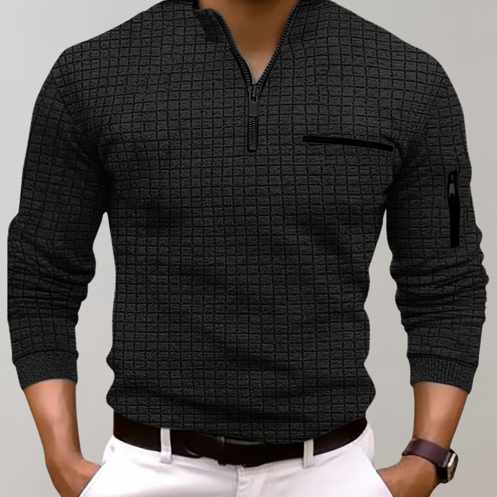 Homme portant un pull noir texturé à col zippé, manches longues, poche poitrine, sur fond gris. Mode masculine élégante et moderne.