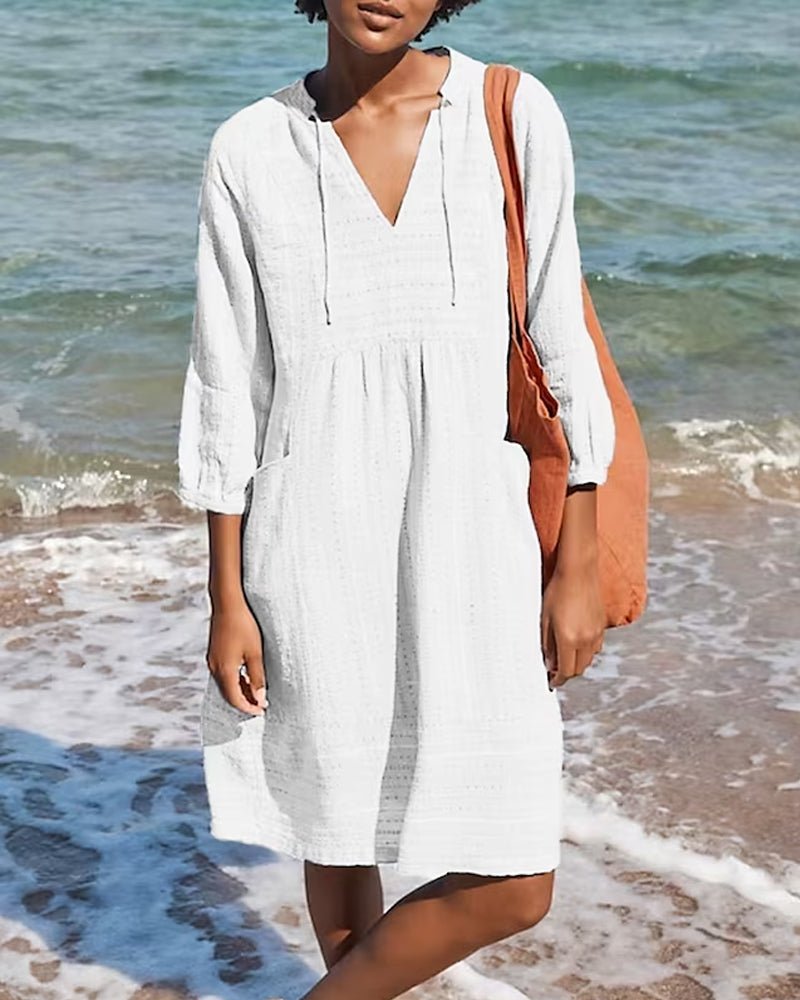 Femme en robe blanche légère sur la plage, portant un sac orange. Mer en arrière-plan. Mode estivale, tenue de plage, style décontracté.