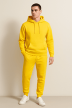 Homme portant un survêtement jaune vif avec capuche, pantalon assorti et baskets blanches, debout contre un mur beige. Mode décontractée tendance.