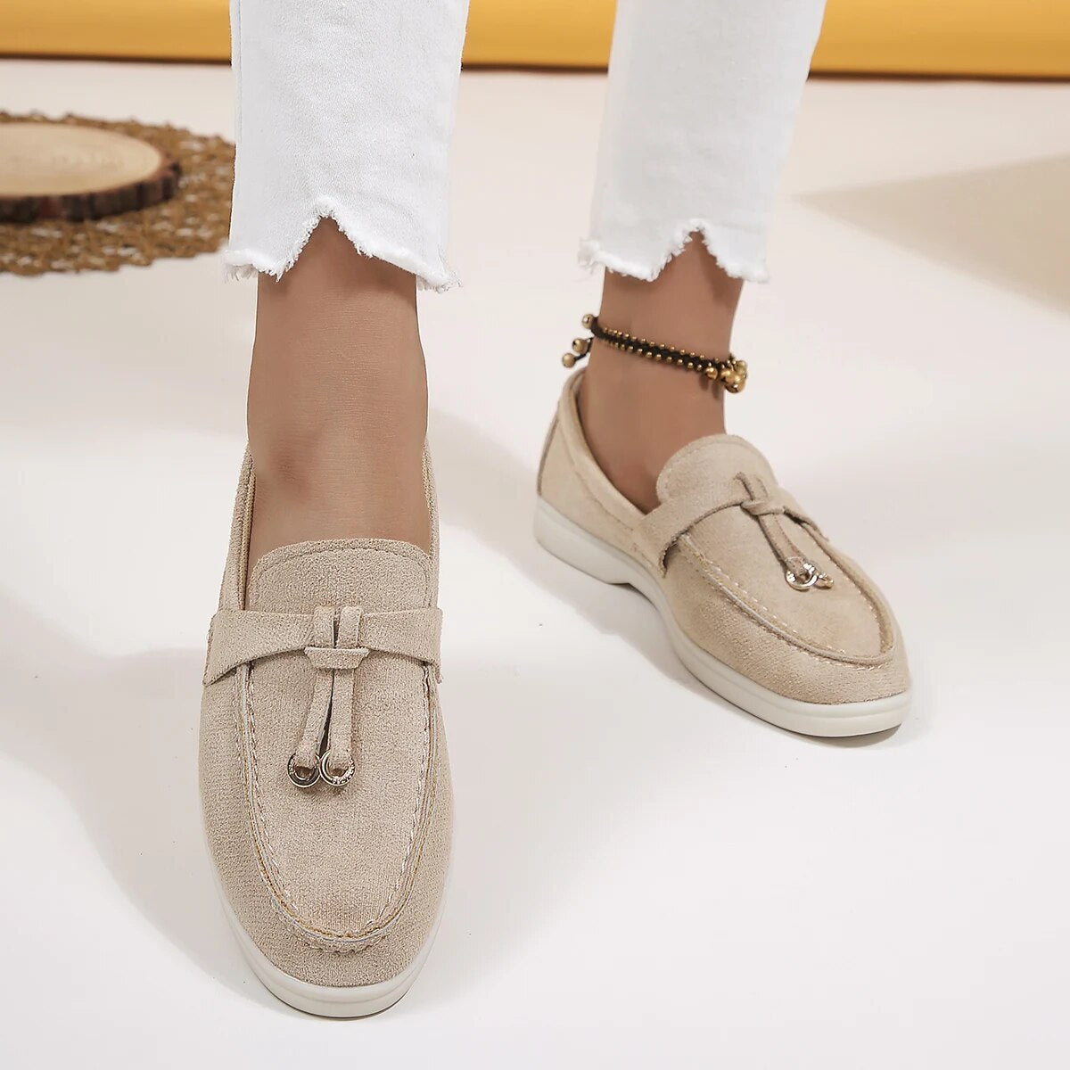 Chaussures mocassins beiges pour femme, en toile, avec détails métalliques, portées avec un pantalon blanc. Mode féminine, style décontracté.