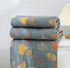 Couverture en lin gris avec motifs de feuilles de ginkgo jaunes et oranges, posée sur une table blanche, à côté d'une lampe moderne.