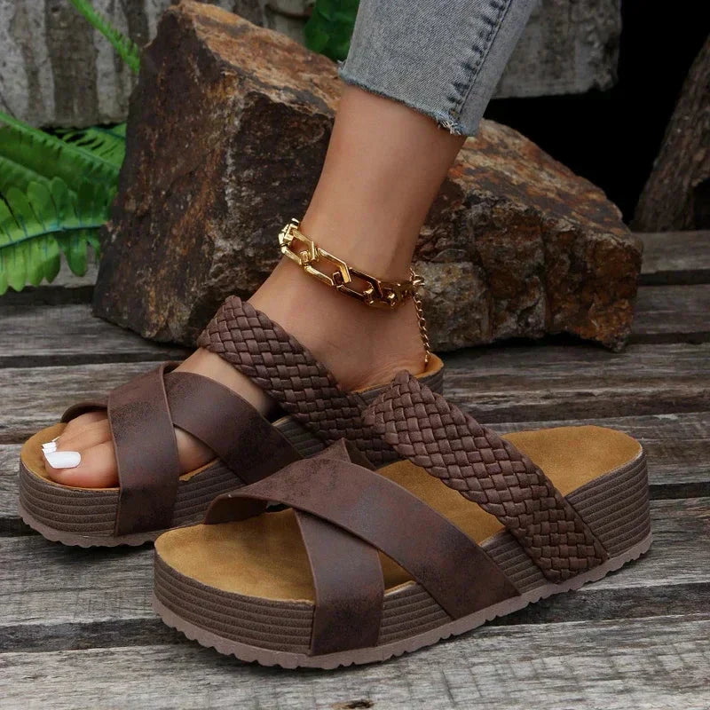 Sandales compensées marron tressées sur fond de bois, portées avec un bracelet de cheville doré. Mode estivale, chaussures tendance, confort.