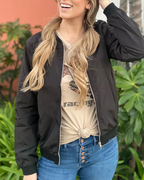 Femme souriante portant un blouson noir, t-shirt beige noué, et jean bleu, posant devant des plantes vertes. Mode décontractée, style urbain.