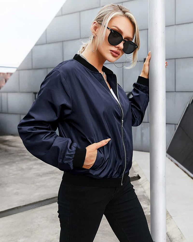Femme en blouson bomber bleu marine, lunettes de soleil noires, posant contre un poteau, style urbain moderne, mode féminine tendance.