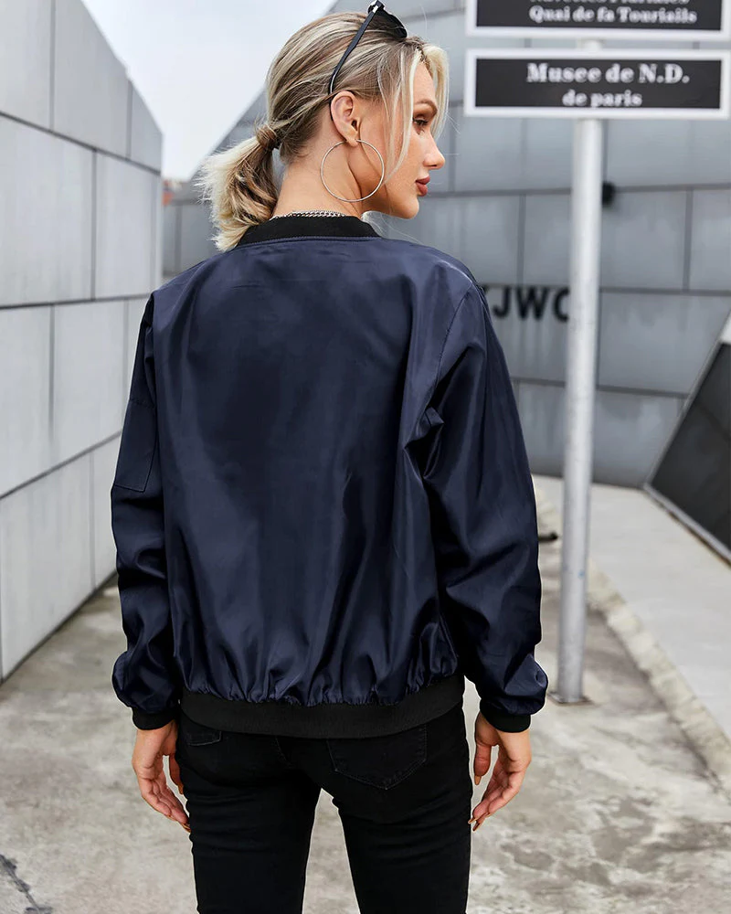 Femme portant un blouson bomber bleu marine, cheveux attachés, lunettes de soleil sur la tête, devant un mur moderne à Paris. Mode urbaine chic.