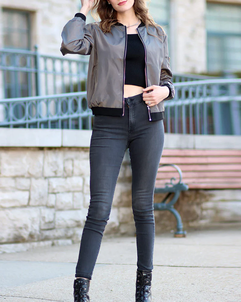 Femme portant un blouson gris, un top noir et un jean slim gris, posant devant un banc en pierre. Mode urbaine, style décontracté, tendance automne.