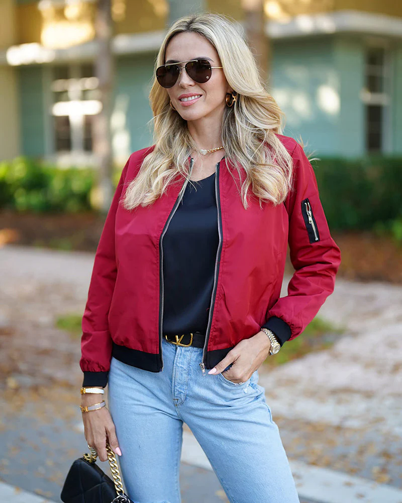 Femme blonde portant une veste rouge, lunettes de soleil et jean bleu, posant à l'extérieur. Mode décontractée, style automnal, look tendance.