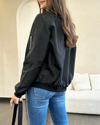 Femme portant un blouson noir élégant, debout dans un salon moderne. Veste légère, jeans ajustés, cheveux longs. Mode décontractée chic.