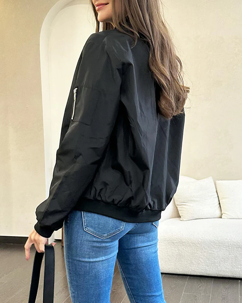 Femme portant un blouson noir élégant, debout dans un salon moderne. Veste légère, jeans ajustés, cheveux longs. Mode décontractée chic.