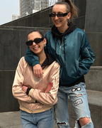 Deux femmes souriantes portant des vestes bomber, lunettes de soleil noires et jeans décontractés, posant devant un mur urbain. Mode urbaine tendance.