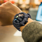 Montre intelligente noire avec écran circulaire affichant des notifications, portée sur un poignet, en arrière-plan flou d'un bureau moderne.