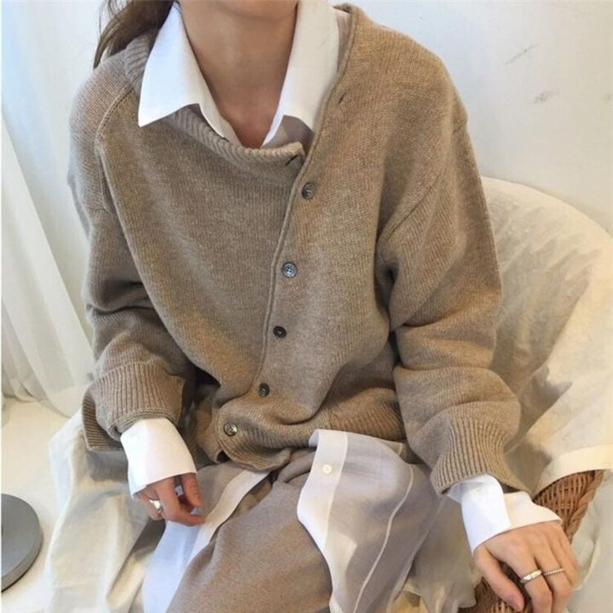 Femme portant un cardigan beige en laine sur une chemise blanche, assise sur une chaise. Mode automne-hiver, style décontracté et élégant.