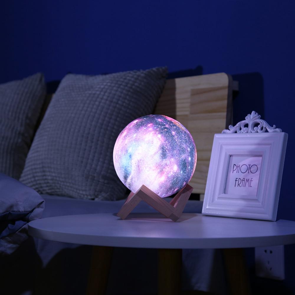 Lampe lune 3D colorée sur table de chevet, éclairant une chambre sombre avec coussins gris et cadre photo blanc. Éclairage décoratif moderne.