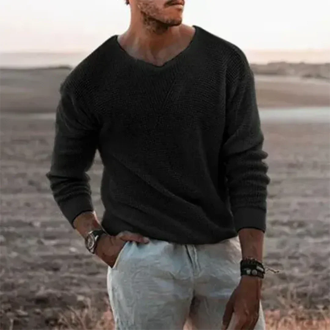 Homme portant un pull noir et un pantalon blanc, debout dans un paysage désertique. Mode masculine, style décontracté, accessoires tendance.