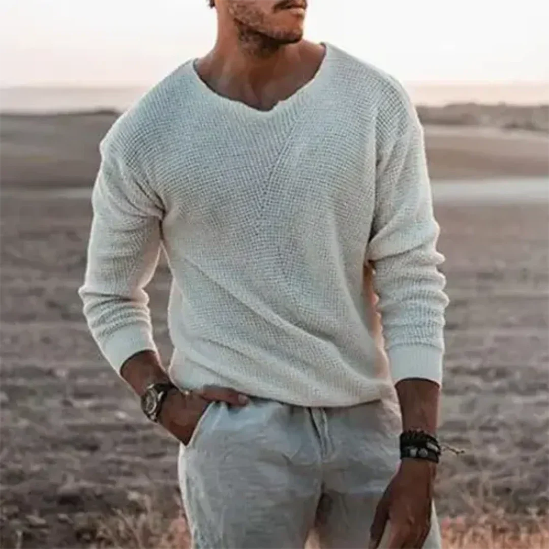 Homme portant un pull blanc texturé et un pantalon beige dans un paysage désertique au coucher du soleil, style décontracté et élégant.