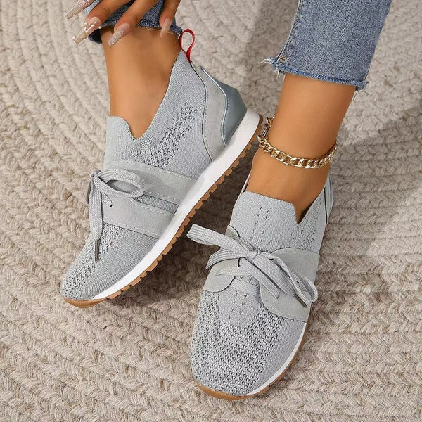 Chaussures de sport grises pour femme avec lacets, semelle blanche et détails en maille. Portées avec un jean et un bracelet doré. Mode décontractée.