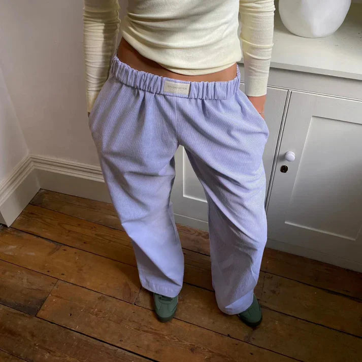 Personne portant un pantalon ample bleu clair en velours côtelé, avec un haut blanc à manches longues, debout sur un sol en bois.