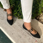 Chaussures noires élégantes avec motif zébré, portées avec un pantalon blanc, posées sur un trottoir en béton près de verdure. Mode féminine chic.