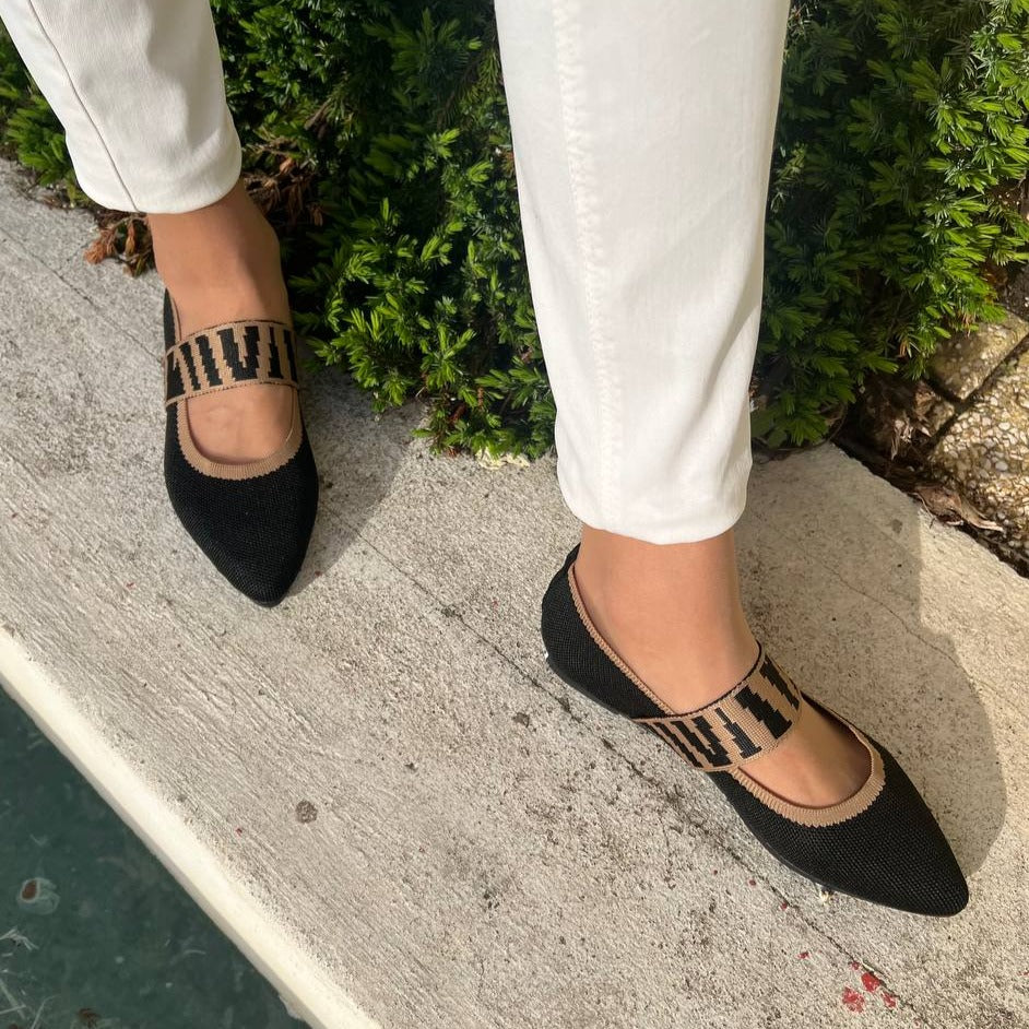 Chaussures noires élégantes avec motif zébré, portées avec un pantalon blanc, posées sur un trottoir en béton près de verdure. Mode féminine chic.