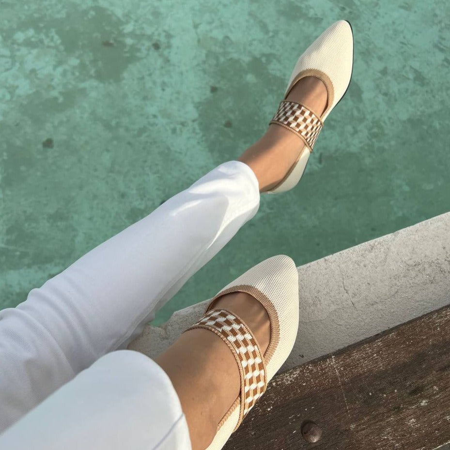 Chaussures plates beiges avec motif à carreaux, portées avec un pantalon blanc, posées près d'une piscine turquoise. Mode estivale élégante.