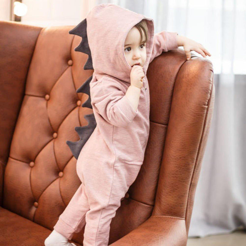 Enfant en combinaison rose avec capuche à écailles de dinosaure, debout sur un canapé en cuir marron. Mode bébé, tenue dinosaure, intérieur maison.