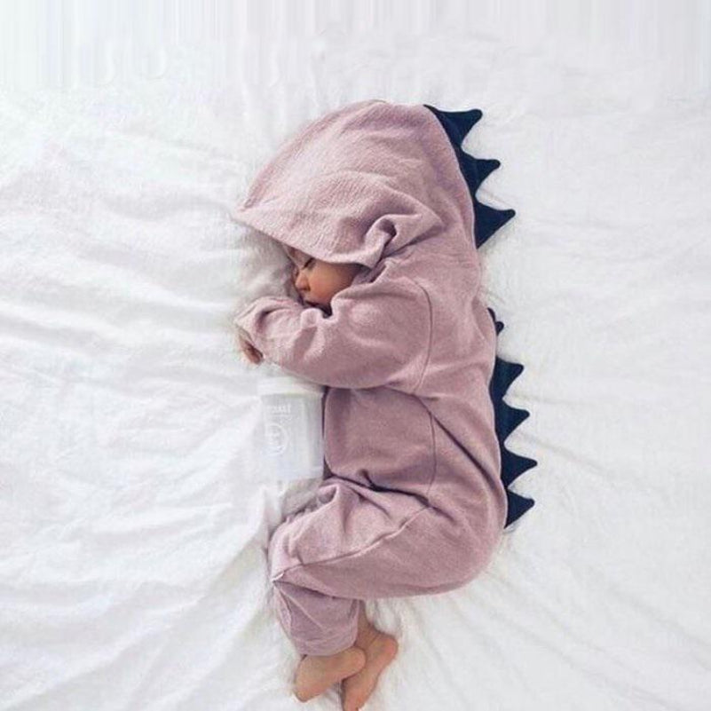 Bébé endormi en pyjama dinosaure rose avec capuche et écailles noires, allongé sur un lit blanc. Pyjama bébé mignon, sommeil enfant paisible.
